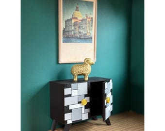 Puede incluir: Un pequeño mueble negro con diseños geométricos grises y blancos y pomos dorados. Una figura de carnero dorado se encuentra encima, con una foto enmarcada de un edificio al fondo. El mueble tiene dos puertas, una abierta.