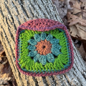 May include: A handmade crochet square with a floral design. The square features a teal and orange flower in the center, surrounded by green leaves. The border is a deep purple color. The square is resting on a piece of wood.