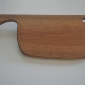 May include: A wooden cutting board with a handle and a rounded shape. The board is made of light brown wood and has a natural grain pattern.