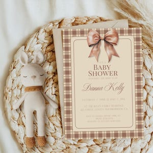 May include: A baby shower invitation with a brown and beige plaid pattern. The invitation features a large brown bow and the text "It's a Baby Shower Honoring the Mom to Be Danna Kelly Saturday April 12, 2025 | 3PM At the Kelly Family Residence 123 Street, Lovely Neighborhood, CA RSVP to Sandra before April 1st Phone 312334 Registry at Pottery Barn Kids"