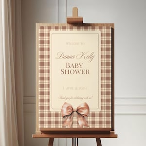 May include: A wooden easel displaying a baby shower sign. The sign features a cream-colored rectangular center with text that reads "Welcome to Danna Kelly Baby Shower" and the date. A large bow is at the bottom.
