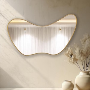 May include: A gold-framed, wavy-shaped mirror hangs on a light beige wall. Two white ceramic vases sit on the floor in front of the mirror, one larger than the other.