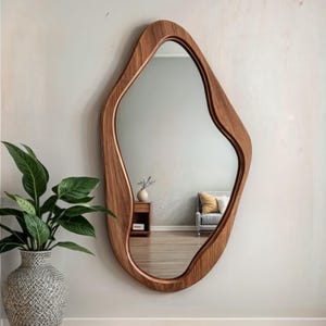 Puede incluir: Un espejo con marco de madera con una forma orgánica y fluida. El espejo está montado en una pared de color beige claro y refleja una sala de estar con un sofá y una mesa auxiliar.
