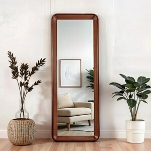 Puede incluir: Un espejo de cuerpo entero con un marco de madera marrón. El espejo está de pie frente a una pared blanca con una planta en maceta a cada lado. El espejo refleja un sillón blanco y una imagen enmarcada.