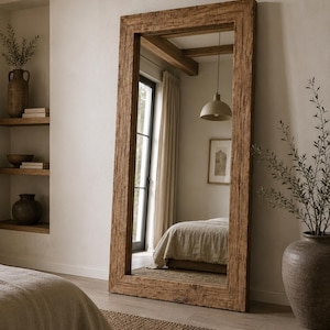 Op de afbeelding: Een hoge, rechthoekige vloerspiegel met een dikke, verweerde bruine houten lijst met een zichtbare houtnerf en een getextureerd oppervlak. De spiegel staat rechtop en reflecteert een slaapkamerinterieur met een bed, een hanglamp en een raam.