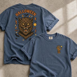 May include: A slate blue t-shirt featuring a large graphic on the back of a snarling werewolf head surrounded by the text HALLOWEEN, bats, candy corn, lightning bolts, and stars. The front chest area displays a small graphic of a furry clawed hand.