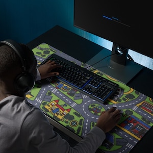 May include: A person using a black keyboard and mouse on a desk. The desk has a colorful road map-themed mousepad. A computer monitor displays a loading screen. The person is wearing headphones.