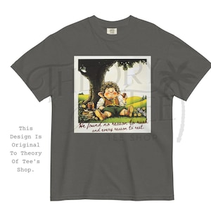 May include: Dark gray t-shirt featuring a framed illustration of a hobbit-like character relaxing under a tree, holding a cup. The image includes the text "He found no reason to rush and every reason to rest." The shirt has the text "This Design Is Original To Theory Of Tee's Shop."