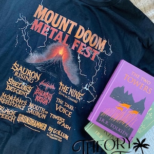 May include: Black t-shirt with orange and white text that reads "MOUNT DOOM METAL FEST" above a graphic of a volcano. Below the text are band names. Two books are next to the shirt, one purple and one green.