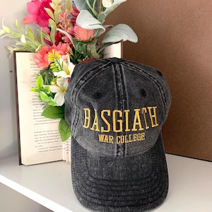 以下が含まれることがあります： 「BASGIATH WAR COLLEGE」の文字が金色の刺繍で施された、黒色のウォッシュ加工ベースボールキャップ。白い棚の上に置かれており、背景には本と花が配置されています。