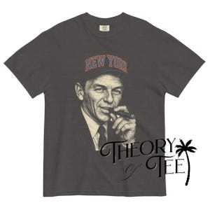 May include: Dark gray t-shirt featuring a black and white portrait of a man wearing a hat with "NEW YORK" in orange lettering. The man is holding a cigar. The shirt has the text "THEORY & TEE" in a cursive font.