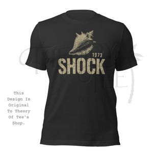 May include: Black t-shirt with a beige conch shell graphic above the word "SHOCK" and the year "1973". The design has a distressed, vintage appearance. The shirt has short sleeves and a crew neck.