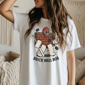 May include: White t-shirt featuring a hockey-themed graphic. The design includes a brick wall, hockey equipment, and the text "BRICK WALL BOB". The shirt is a casual, everyday wear item.