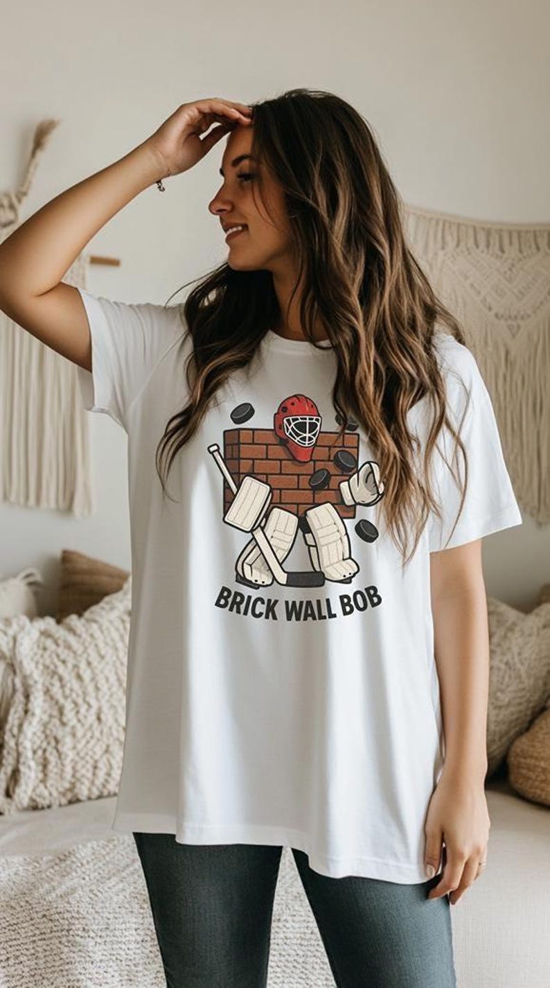 May include: White t-shirt featuring a hockey-themed graphic. The design includes a brick wall, hockey equipment, and the text "BRICK WALL BOB". The shirt is a casual, everyday wear item.