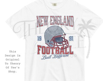 以下が含まれることがあります： 白のTシャツ。赤と青で「NEW ENGLAND FOOTBALL」の文字と、グレーのアメリカンフットボールのヘルメットのグラフィックが特徴です。「19」と「91」の数字がヘルメットの両側にあります。「FOOTBALL」の下には「Built Different」の文字があります。