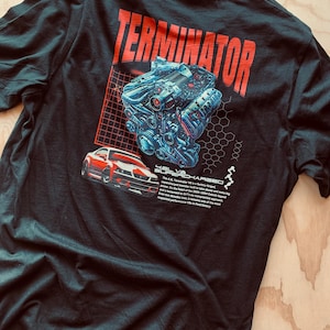 Puede incluir: Camiseta negra con un diseño gráfico rojo y azul. El diseño incluye la palabra "TERMINATOR" en rojo, una ilustración de motor azul y un coche rojo y blanco. El texto debajo del coche dice "4.6L SUPERCHARGED".