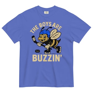 Puede incluir: Camiseta azul con una abeja de dibujos animados jugando al hockey. La abeja lleva un casco azul y patines, sosteniendo un palo de hockey. El texto en la camiseta dice "THE BOYS ARE BUZZIN'" en un estilo vintage.