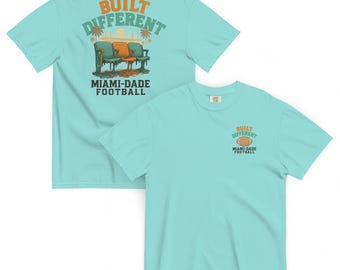 Könnte beinhalten: Zwei hellblaue T-Shirts. Eines zeigt eine Grafik von Stadionsitzen mit dem Text "BUILT DIFFERENT" und "MIAMI-DADE FOOTBALL". Das andere Shirt hat eine kleinere Grafik eines Fußballs und denselben Text. Die Shirts sind aus weichem Material.