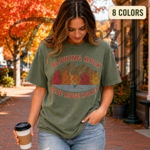 May include: An olive green short-sleeve t-shirt featuring a graphic of mountains and autumn trees with the text "BLOWING ROCK" and "BLUE RIDGE HOCKEY" in red. A small figure is depicted skating on a frozen surface in the center of the design.