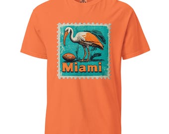 Könnte beinhalten: Orangefarbenes T-Shirt mit einer Vintage-Illustration eines weißen Ibis-Vogels mit orangefarbenem Schnabel und Beinen, der auf einem grünen Hintergrund mit einem Fußball und dem Wort "Miami" in einem blauen und orangefarbenen Stempel steht.