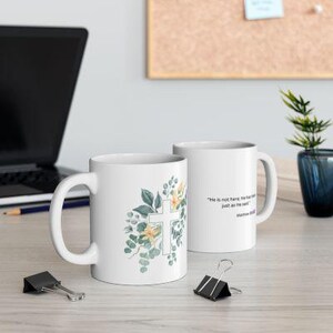 Könnte beinhalten: Weiße Keramiktasse mit Blumenmuster und einem weißen Kreuz. Die Tasse trägt den Spruch "Er ist nicht hier, er ist auferstanden, wie er gesagt hat." aus Matthäus 28,6.