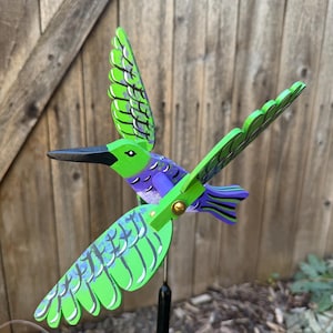 May include: A wooden hummingbird garden spinner featuring vibrant green and purple painted feathers with black detailing. The bird has a long black beak and is mounted on a thin black metal rod, designed to rotate with the wind.