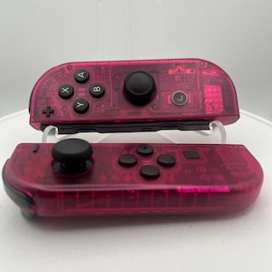 May include: A pair of pink transparent Nintendo Switch Joy-Con controllers. The controllers have black buttons and a black analog stick.