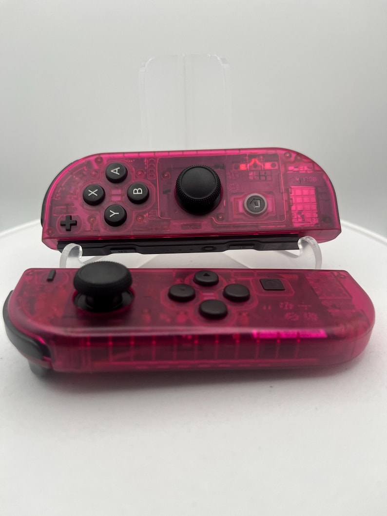 May include: A pair of pink transparent Nintendo Switch Joy-Con controllers. The controllers have black buttons and a black analog stick.