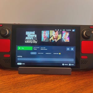 May include: A black handheld gaming console with red custom buttons, a red directional pad, and red trackpads. The screen displays the Grand Theft Auto IV and Episodes from Liberty City game menu, showing a play button and game statistics.