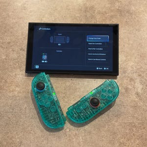 May include: A black handheld gaming console displaying a controller settings menu on its screen, accompanied by two translucent teal wireless controllers featuring black joysticks and buttons, positioned below the console screen.