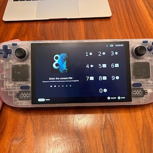 May include: A handheld gaming console with a transparent shell, featuring a black screen displaying a PIN entry interface with numbers 0 through 9, the text Enter the current PIN, and various controller button icons including L1, L2, R1, R2, and Y.