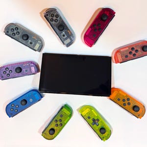May include: A black rectangular gaming console screen surrounded by eight individual game controllers in various translucent colors including gray, pink, red, orange, yellow-green, bright green, blue, and purple, each featuring black buttons and joysticks.