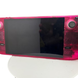 May include: A transparent pink handheld gaming console with a black screen and black buttons. The console has a D-pad, analog sticks, and action buttons. The word "STEAM" is visible on the console.