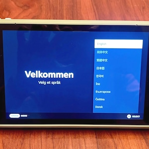 May include: A sage green handheld gaming console with white buttons and joysticks. The screen displays a blue menu with the text Velkommen and a list of languages for selection. The device features a directional pad, two analog sticks, and trackpads.