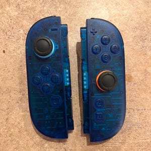 May include: Two transparent blue game controllers with black and orange buttons. The controllers are split, showing the internal components. The left controller has a directional pad and a black joystick. The right controller has action buttons and a black joystick.