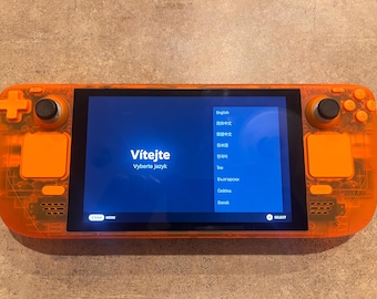 May include: A handheld gaming console with a translucent orange shell. The screen displays a blue menu with the text Vítejte Vyberte jazyk, alongside a list of languages. It features two black analog sticks, a directional pad, and orange buttons.