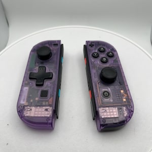 May include: A pair of purple translucent Nintendo Switch Joy-Con controllers. The controllers have black buttons and a black d-pad. The controllers are separated and facing the camera.