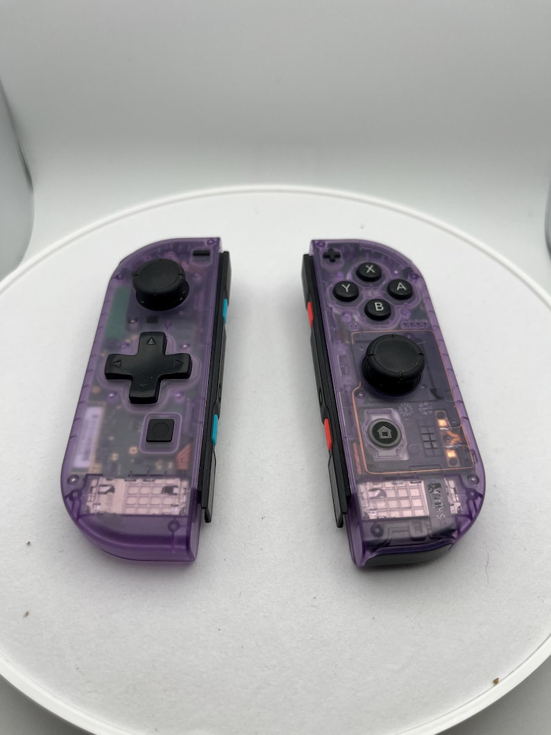 May include: A pair of purple translucent Nintendo Switch Joy-Con controllers. The controllers have black buttons and a black d-pad. The controllers are separated and facing the camera.