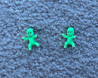 Op de afbeelding: Een paar oorbellen met groene, in het donker lichtgevende babyfiguurtjes. De oorbellen hebben zilveren haakjes.