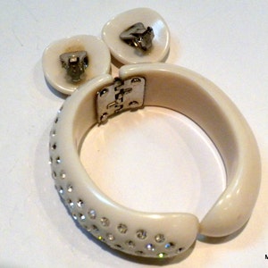 May include: Ivory-colored vintage jewelry set featuring a hinged bangle bracelet and matching clip-on earrings. The bracelet is adorned with sparkling rhinestones along the front. The earrings and bracelet have silver-tone metal hardware.