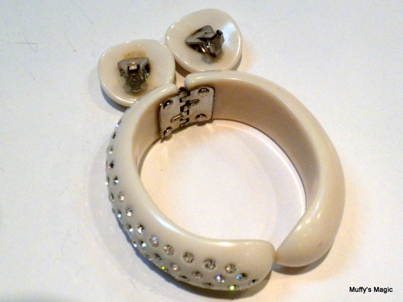 May include: Ivory-colored vintage jewelry set featuring a hinged bangle bracelet and matching clip-on earrings. The bracelet is adorned with sparkling rhinestones along the front. The earrings and bracelet have silver-tone metal hardware.