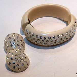 May include: Ivory-colored jewelry set featuring a hinged bangle bracelet and matching clip-on earrings. The bracelet and earrings are adorned with numerous sparkling rhinestones, creating a glamorous and eye-catching design. A classic accessory set.