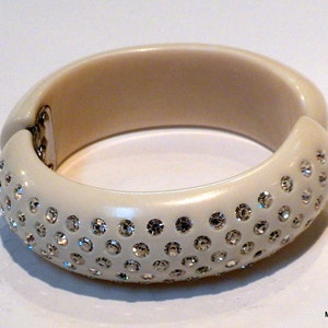 May include: Ivory-colored hinged bangle bracelet. The bracelet is adorned with numerous sparkling rhinestones on the outer surface. The clasp is silver-toned. A stylish accessory for any outfit.