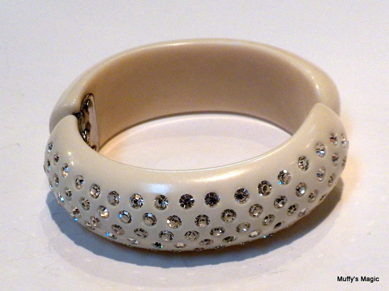 May include: Ivory-colored hinged bangle bracelet. The bracelet is adorned with numerous sparkling rhinestones on the outer surface. The clasp is silver-toned. A stylish accessory for any outfit.