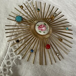 May include: A gold-toned sunburst brooch with a central floral design and radiating spokes. The brooch is adorned with various colorful gemstones, including turquoise, red, and clear crystals. The brooch is set against a white, textured background.