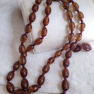 May include: A vintage necklace featuring three strands of large, oval amber-colored beads. The beads are separated by small, dark red beads. The necklace is fastened with a decorative clasp with a purple stone.