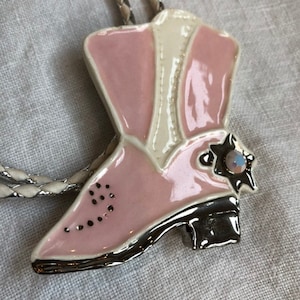 May include: A pink and white ceramic cowboy boot pendant with a silver-tone braided cord. The boot features a star-shaped embellishment with a small iridescent stone and a black heel. The boot is approximately 2 inches in length.