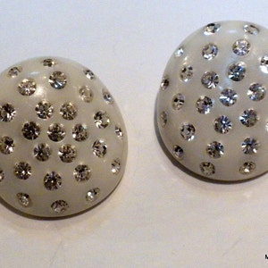 May include: Two off-white, oval-shaped earrings. Each earring is covered with numerous, sparkling, clear rhinestones. The earrings have a smooth, glossy finish and are set against a white background. The earrings are a type of jewelry.