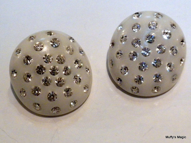 May include: Two off-white, oval-shaped earrings. Each earring is covered with numerous, sparkling, clear rhinestones. The earrings have a smooth, glossy finish and are set against a white background. The earrings are a type of jewelry.