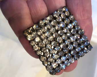 May include: A rectangular, vintage brooch densely covered with small, clear rhinestones. The rhinestones are set in a dark metal base, creating a sparkling, textured surface. The brooch is held in a hand.